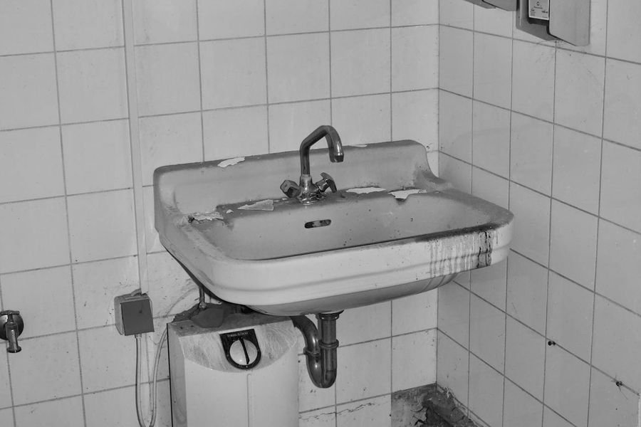 Derelict tiled washroom with a stained basin, an old water heater below it and debris on the floor