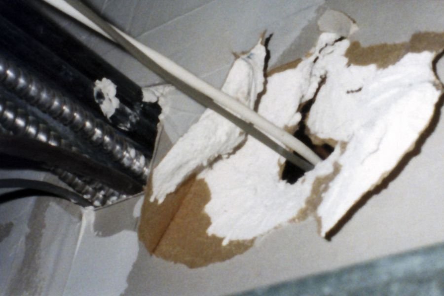 Cables passing through a torn hole in a fire-rated plasterboard wall above a suspended ceiling, with no seal of any kind