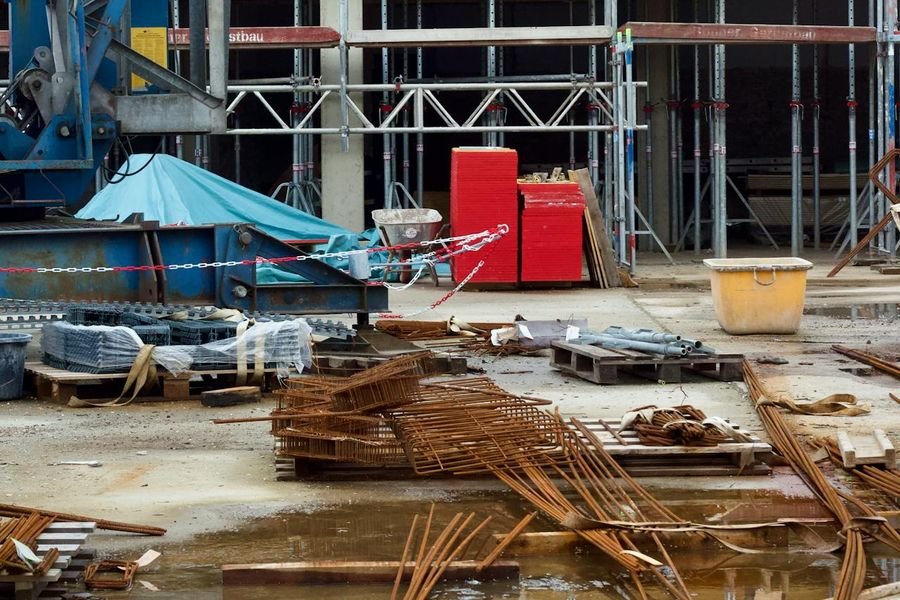 Construction site with stacked steel reinforcement, materials on pallets and puddles in front of a scaffolded building