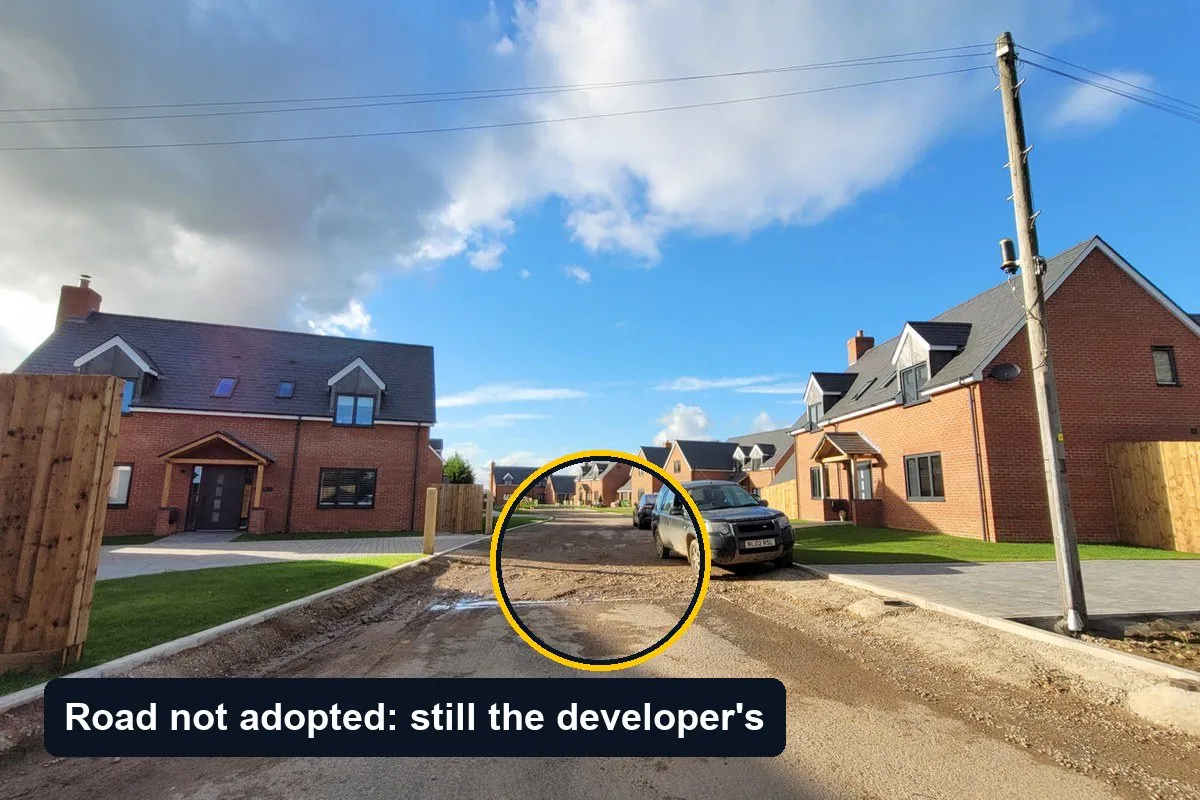 A new build housing street in England with brick houses, a timber fence and an unfinished road surface, circled, with the label Road not adopted: still the developer's